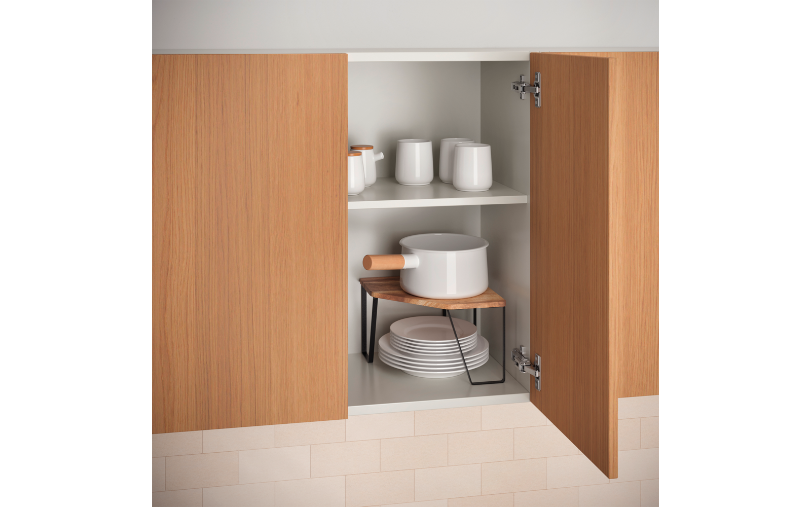 Offener Holzschrank mit zwei Türen und zwei Regalfächern. Oben drei weiße Tassen und zwei kleine weiße Gläser/Dosen. Unten ein weißer Topf mit Holzgriff auf einem kleinen Holzbrett mit schwarzen Metallbeinen, darunter ein Stapel weißer Teller