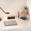 Weißer Schreibtisch vor Wand mit geschlossenem silbernem Laptop, schwarzer Digitaluhr, flacher Schreibtischlampe, kleinem Holz-Eckregal mit Taschentuchbox, Stiftebecher mit Schere und Stiften und darunter Notizbuch und Smartphone