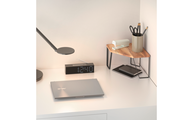 Weißer Schreibtisch vor Wand mit geschlossenem silbernem Laptop, schwarzer Digitaluhr, flacher Schreibtischlampe, kleinem Holz-Eckregal mit Taschentuchbox, Stiftebecher mit Schere und Stiften und darunter Notizbuch und Smartphone