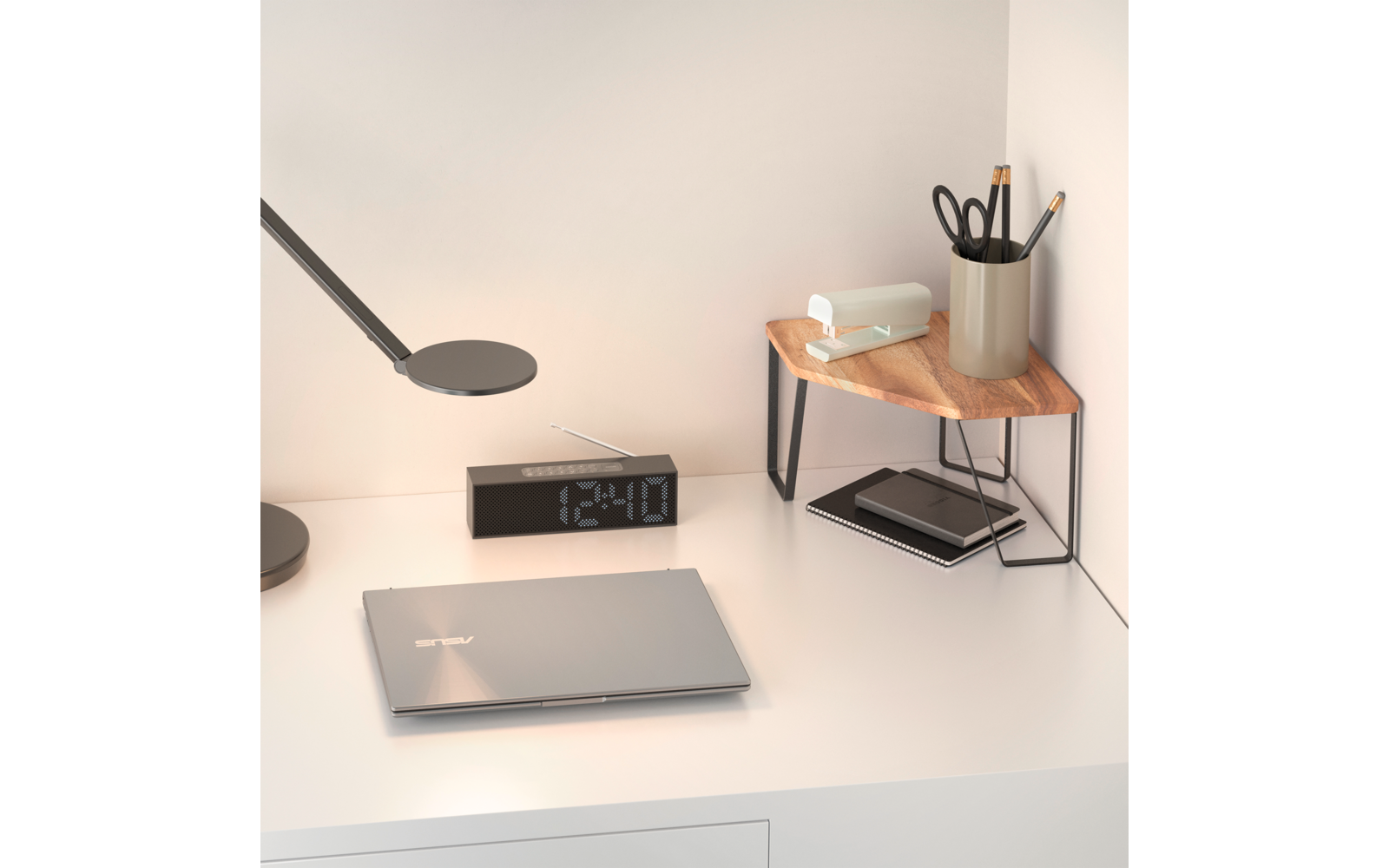 Weißer Schreibtisch vor Wand mit geschlossenem silbernem Laptop, schwarzer Digitaluhr, flacher Schreibtischlampe, kleinem Holz-Eckregal mit Taschentuchbox, Stiftebecher mit Schere und Stiften und darunter Notizbuch und Smartphone
