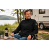 Een man zit in een campingstoel aan een tafel; op de tafel een lantaarn, op de achtergrond een camper en een meer