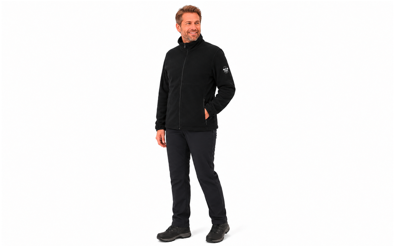 Een man staat voor een witte achtergrond, draagt een zwarte fleecejas, zwarte broek en zwarte schoenen, rechterhand in de jaszak, glimlacht