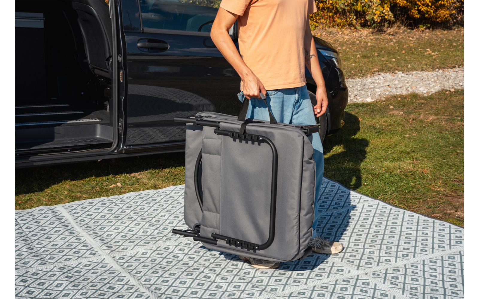 Persona (testa non visibile) con maglietta arancione e pantaloncini blu tiene una borsa grigia piegata con manico e telaio neri; in piedi su un tappeto a motivo accanto a un veicolo nero con portiera aperta, erba e alberi sullo sfondo