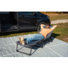 Uomo con T‑shirt arancione e jeans sdraiato su una chaise longue pieghevole, mani dietro la testa, occhiali da sole, gambe distese con i piedi incrociati; accanto a lui un furgone nero con sportello scorrevole aperto; la sdraio è su un tappeto da esterno a motivo, erba e ghiaia sullo sfondo