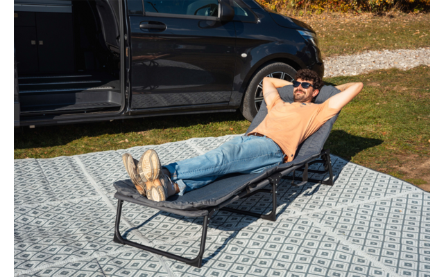 Uomo con T‑shirt arancione e jeans sdraiato su una chaise longue pieghevole, mani dietro la testa, occhiali da sole, gambe distese con i piedi incrociati; accanto a lui un furgone nero con sportello scorrevole aperto; la sdraio è su un tappeto da esterno a motivo, erba e ghiaia sullo sfondo