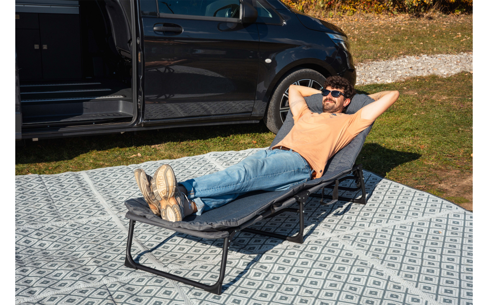 Uomo con T‑shirt arancione e jeans sdraiato su una chaise longue pieghevole, mani dietro la testa, occhiali da sole, gambe distese con i piedi incrociati; accanto a lui un furgone nero con sportello scorrevole aperto; la sdraio è su un tappeto da esterno a motivo, erba e ghiaia sullo sfondo