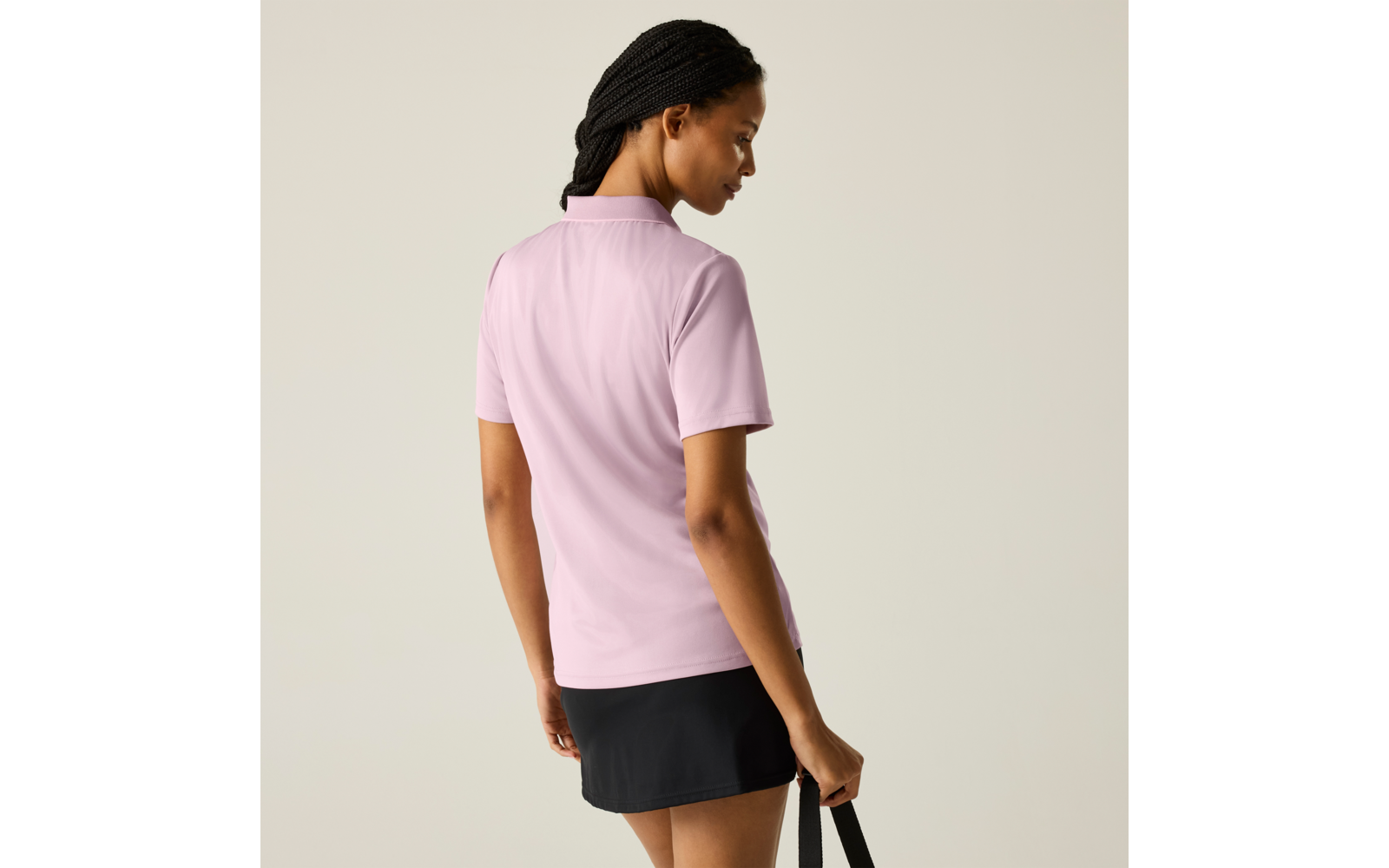 Person von hinten, trägt hellrosa kurzärmeliges Poloshirt und schwarzen Rock, geflochtene Haare, hält einen schwarzen Trageriemen; einfarbiger heller Hintergrund