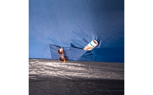 Braune Sonnenbrille und weiße Flasche mit buntem Etikett in einer aufgesetzten Tasche aus blauem Kunststoff, darunter schwarze Kunststofffläche