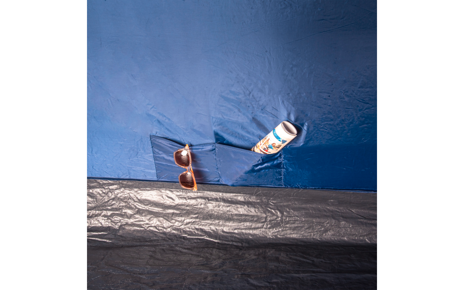 Braune Sonnenbrille und weiße Flasche mit buntem Etikett in einer aufgesetzten Tasche aus blauem Kunststoff, darunter schwarze Kunststofffläche