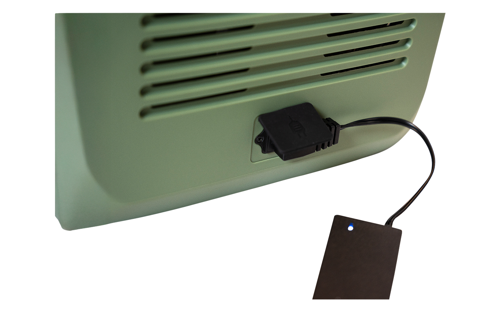 Grünes Gerät mit Lüftungsschlitzen und schwarzem Kabel, das zu einem kleinen schwarzen rechteckigen Gerät mit blauem Leuchtpunkt auf weißer Oberfläche führt