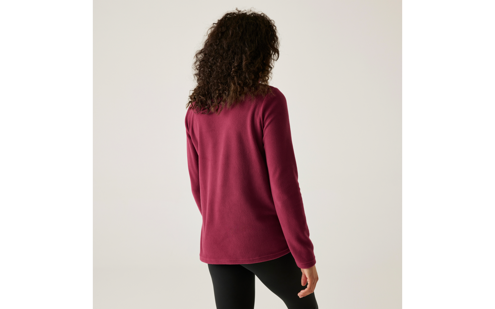 Personne aux cheveux bouclés et foncés vue de dos, portant un pull bordeaux et des leggings noirs sur un fond clair