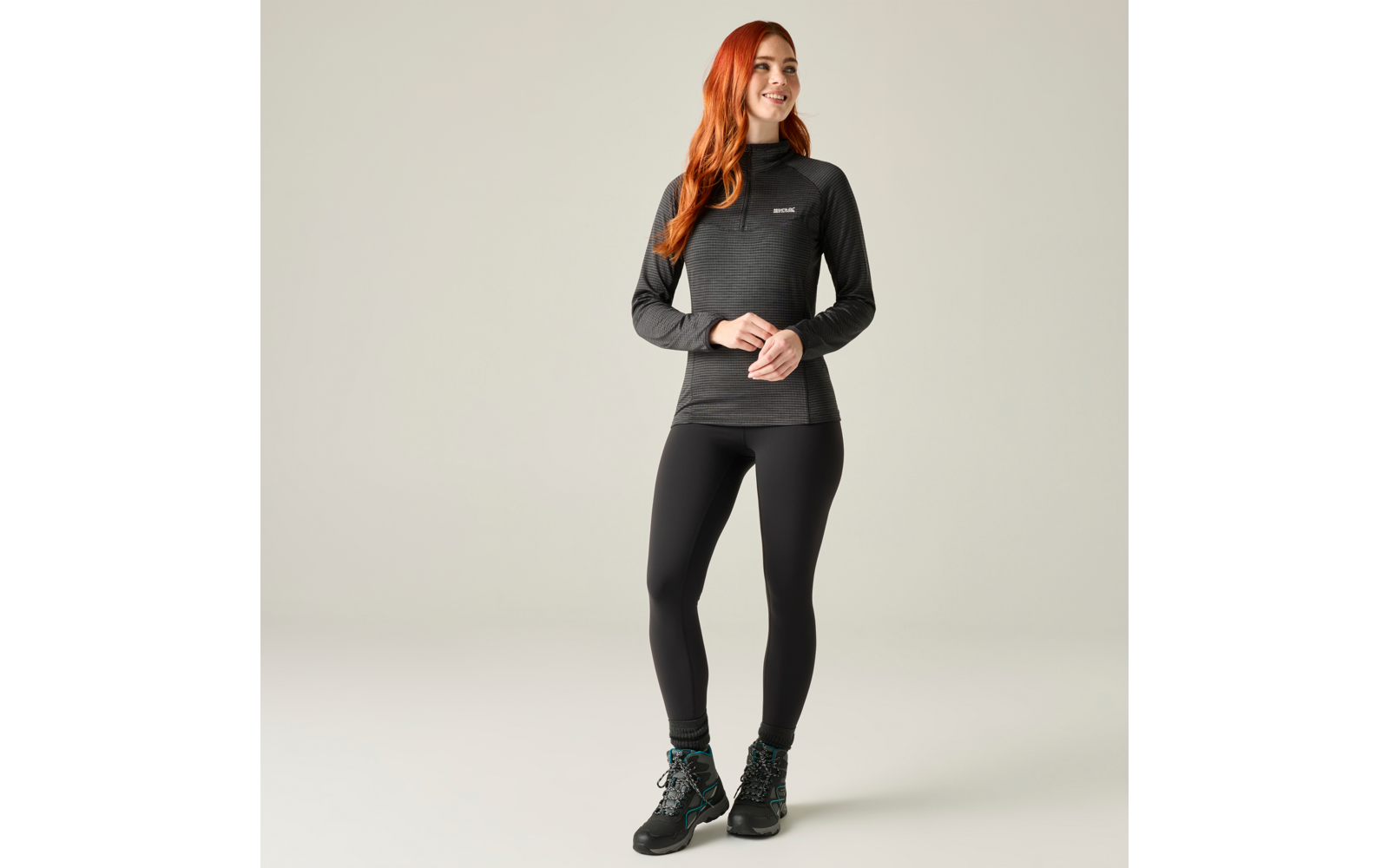 Persoon met lang rood haar staand voor een effen achtergrond, draagt een donker shirt met lange mouwen, zwarte legging en wandelschoenen, handen gevouwen voor zich, kijkt iets naar rechts
