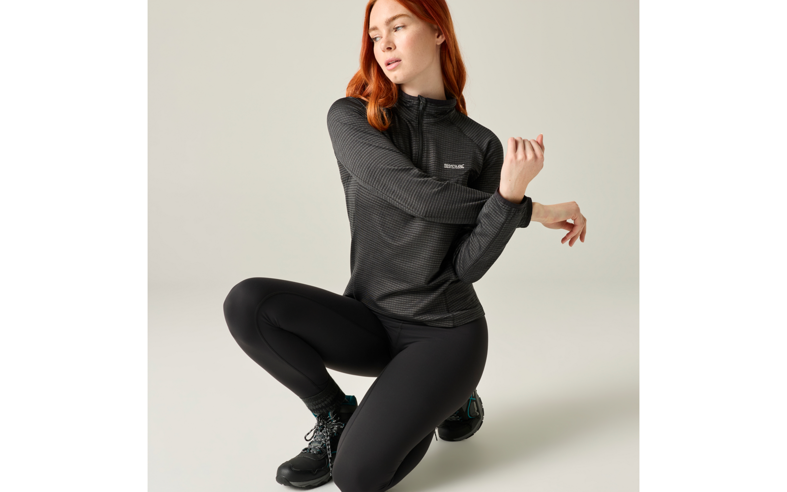 Roodharige vrouw in zwarte sportkleding knielt op één knie voor een lichte achtergrond en trekt aan haar mouw