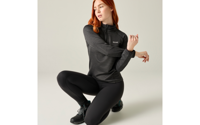 Roodharige vrouw in zwarte sportkleding knielt op één knie voor een lichte achtergrond en trekt aan haar mouw
