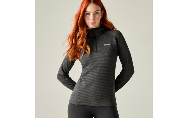 Vrouw met lang rood haar, donkergrijze sporttrui met korte rits en zwarte legging, handen achter de rug, lichte achtergrond
