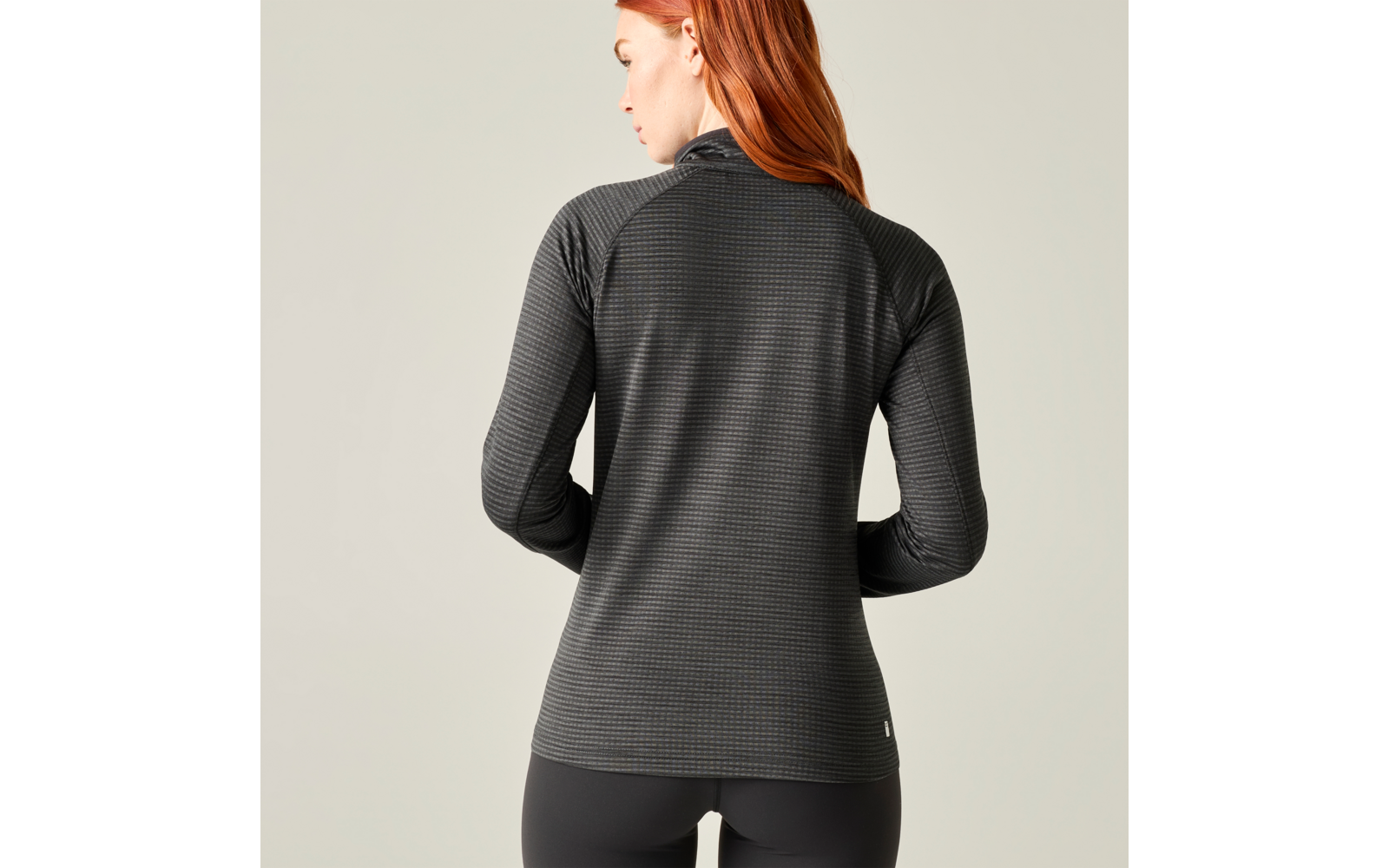 Achteraanzicht van een persoon met rood haar die een donkergrijze, gestructureerde lange mouw top en zwarte leggings draagt voor een licht, neutrale achtergrond