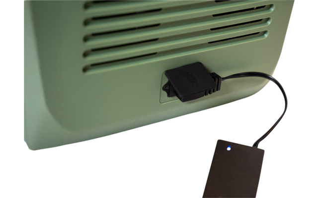 Grünes Gerät mit Lüftungsschlitzen und schwarzem Kabel, das zu einem kleinen schwarzen rechteckigen Gerät mit blauem Leuchtpunkt auf weißer Oberfläche führt