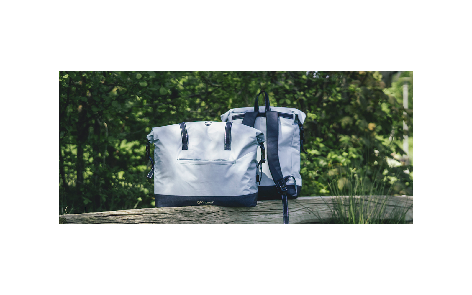 Due borse grigio chiaro (a sinistra una borsa a tracolla, a destra uno zaino) su una ringhiera di legno davanti a cespugli verdi e folti