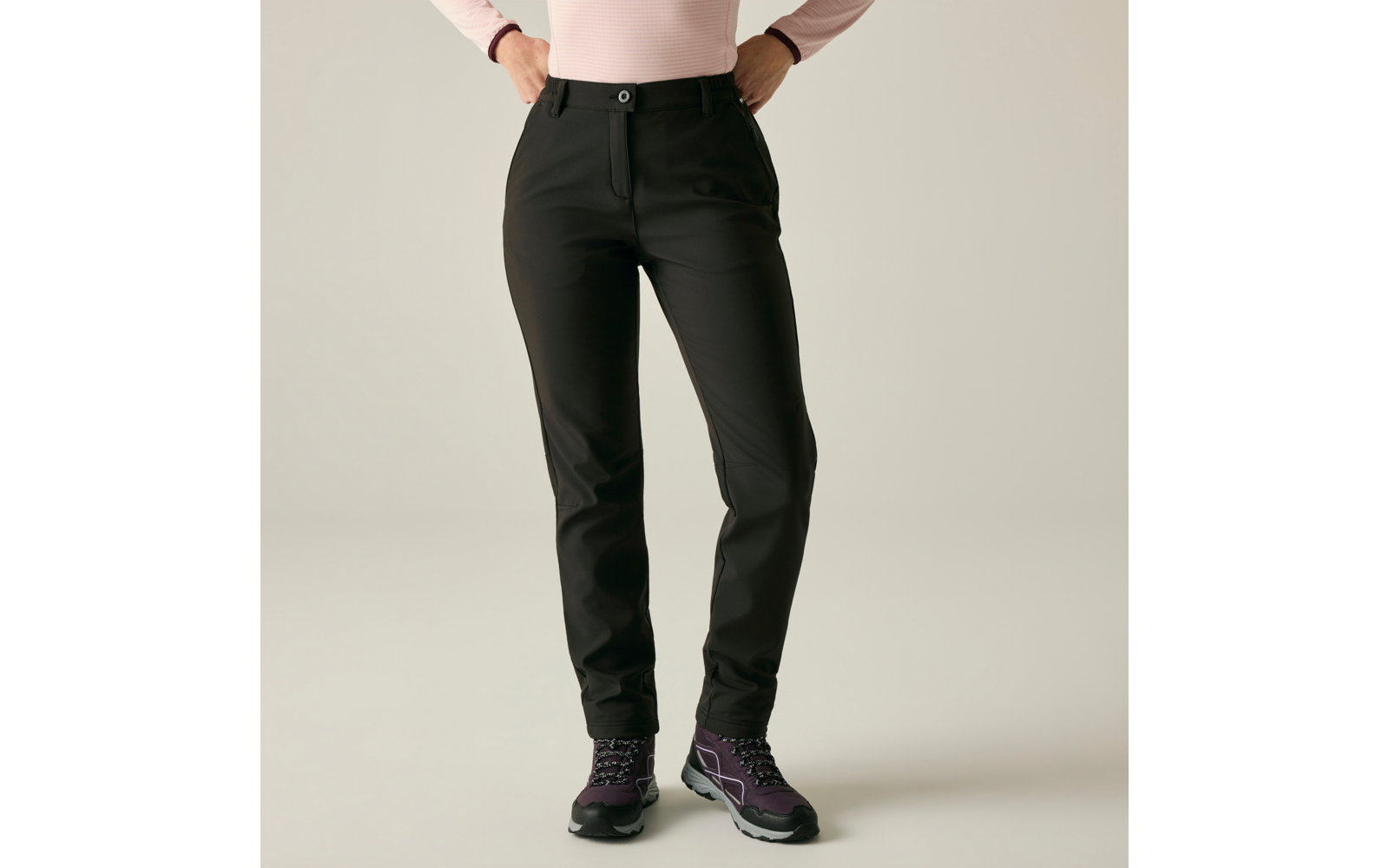 Onderlichaam en heupen van een persoon met zwarte broek en roze longsleeve, handen in de heupen, paarse wandelschoenen, lichte achtergrond