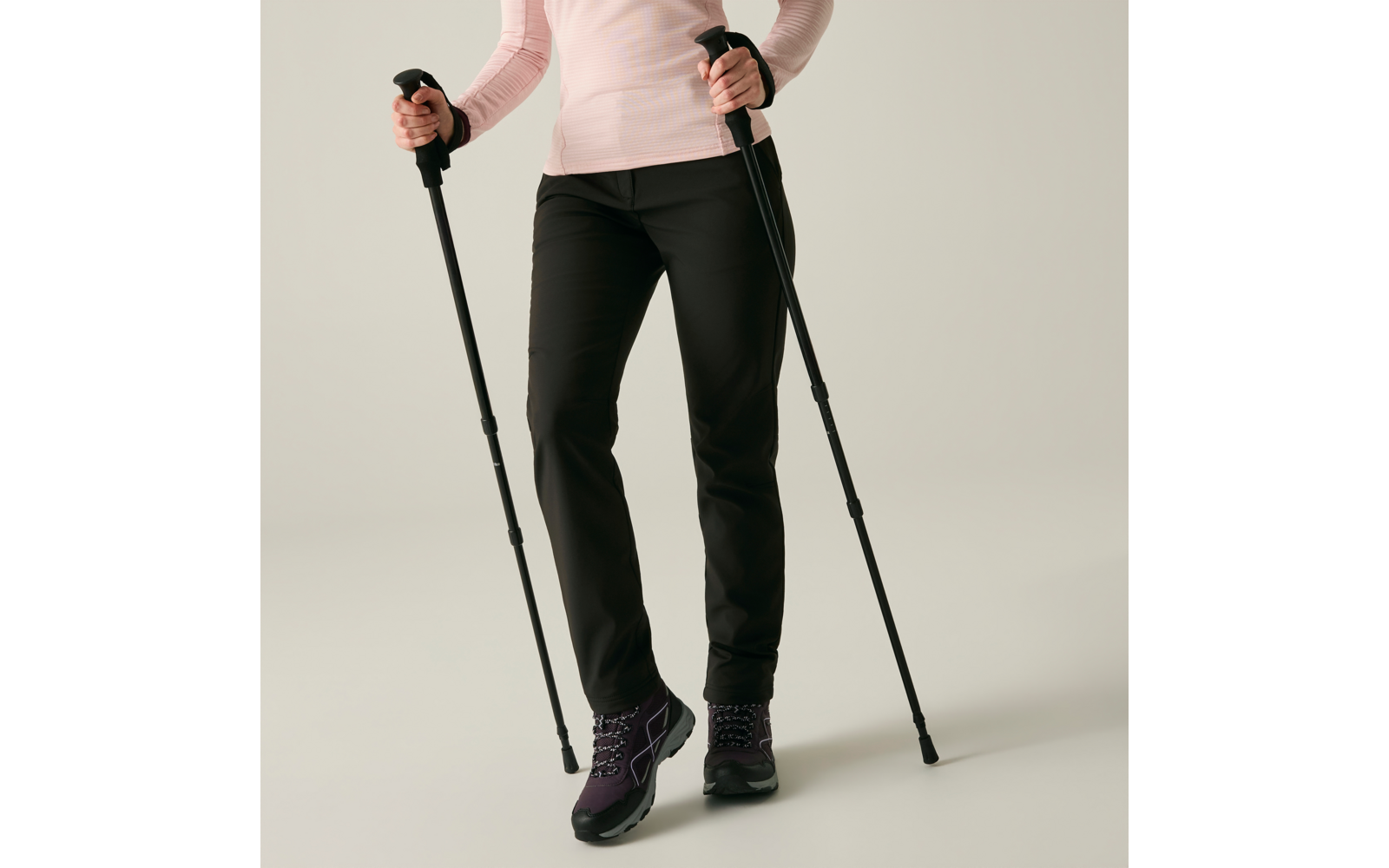 Onderlichaam en handen van een persoon die twee wandelstokken vasthoudt; roze shirt met lange mouwen, zwarte broek, paarse wandelschoenen, lichte effen achtergrond
