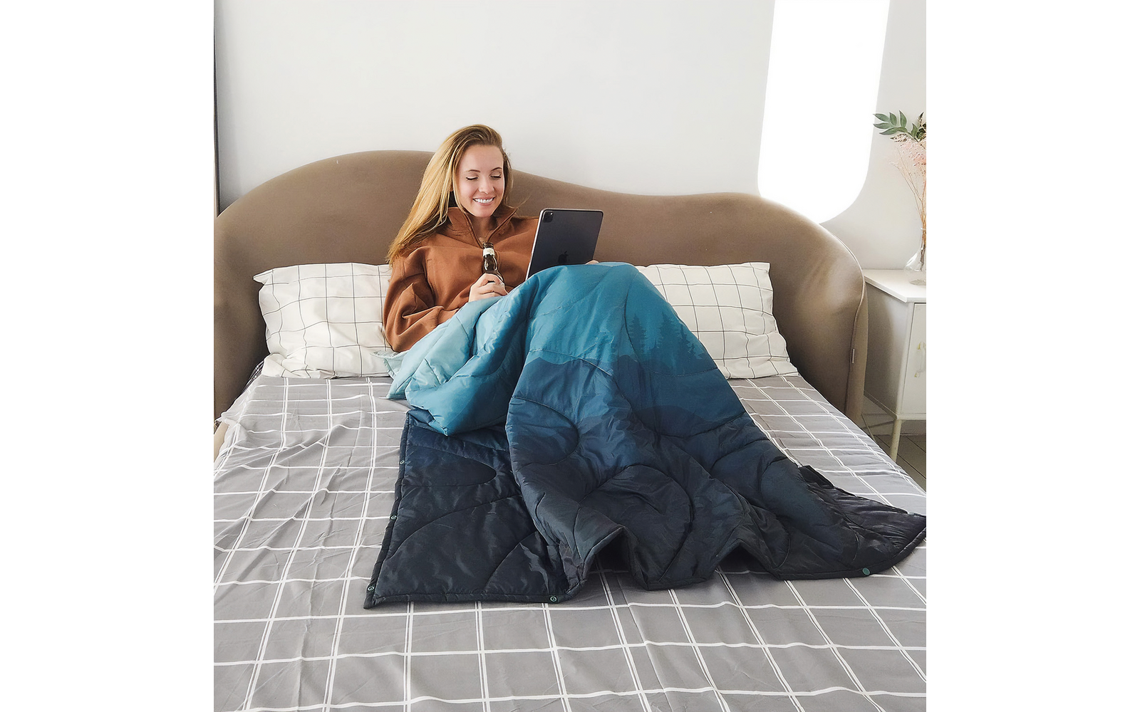 Frau liegt mit einem Tablet in der Hand unter einer blauen Decke auf einem Bett
