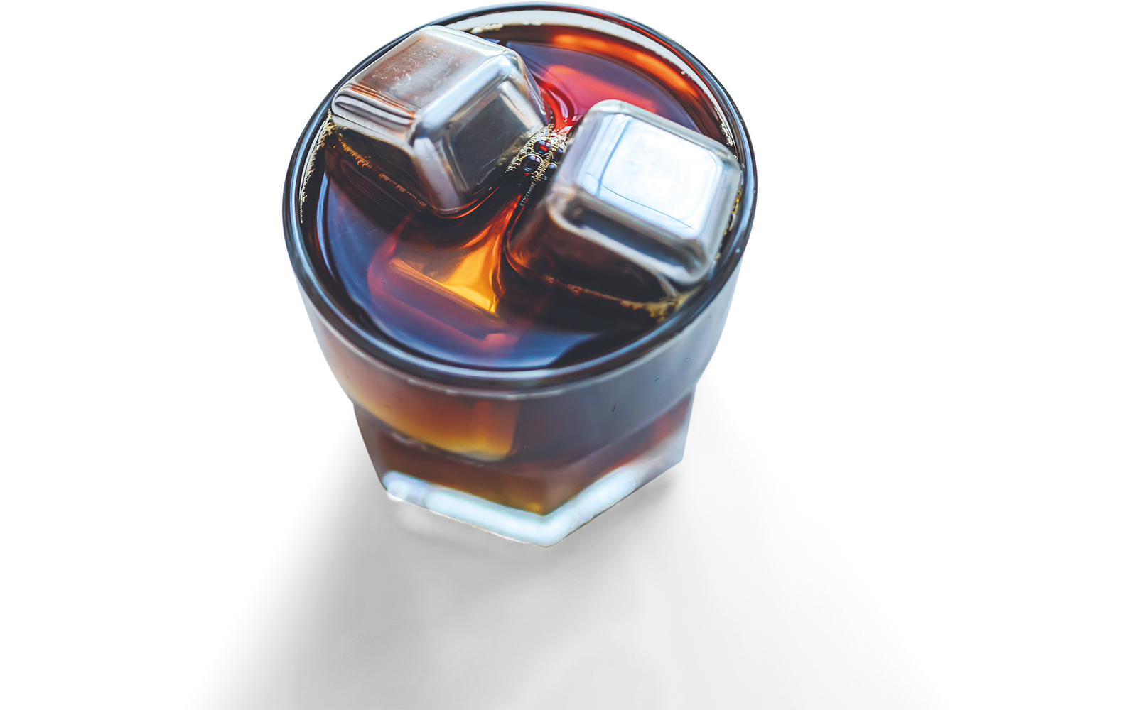 Kurzes facettiertes Glas mit dunkelbraunem Getränk und zwei metallischen Eiswürfeln auf weißem Hintergrund