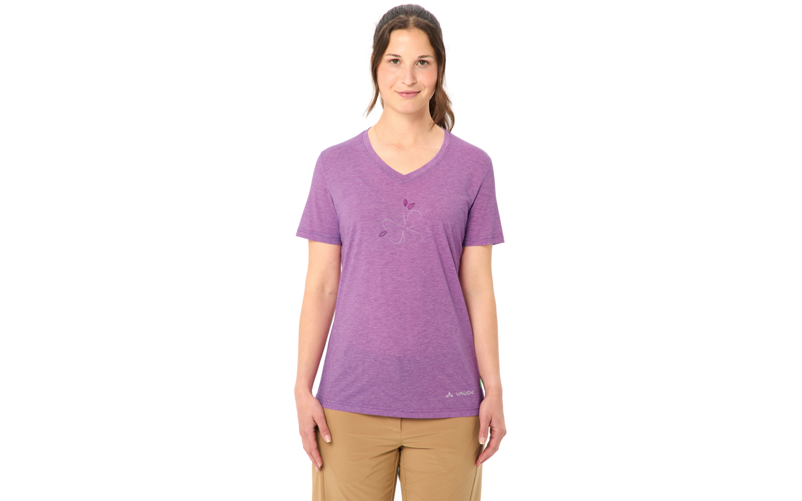 Frau steht frontal vor weißem Hintergrund, trägt lila V‑Ausschnitt-T-Shirt mit kleinem floralem Druck, beige Hose, Haare zurückgebunden, Hände seitlich