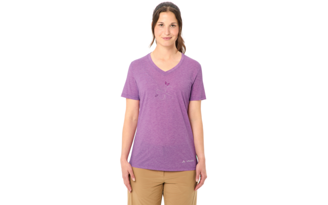 Frau steht frontal vor weißem Hintergrund, trägt lila V‑Ausschnitt-T-Shirt mit kleinem floralem Druck, beige Hose, Haare zurückgebunden, Hände seitlich