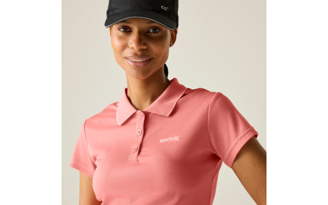 Person mit schwarzer Baseballkappe und rosa Poloshirt mit kleinem weißem Logo auf der linken Brust, lächelnd, Haare nach hinten gebunden, neutraler beiger Hintergrund