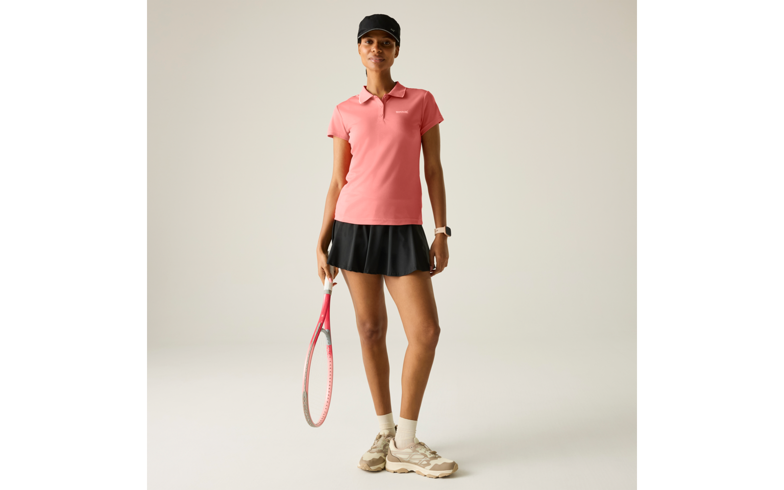 Person in rosa Poloshirt, schwarzem Tennisrock und schwarzer Kappe, hält einen Tennisschläger; trägt weiße Socken, beige Sportschuhe und eine Armbanduhr