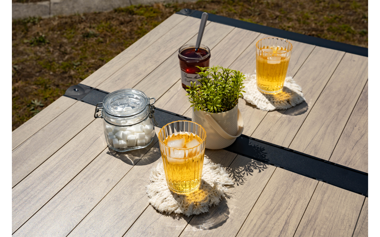 Holztisch im Freien mit zwei Gläsern gelbem Getränk und Eiswürfeln auf gehäkelten Untersetzern, Glas mit Zuckerwürfeln, Glas mit dunkler Marmelade und Löffel, kleiner Topf mit grüner Pflanze, Rasen im Hintergrund