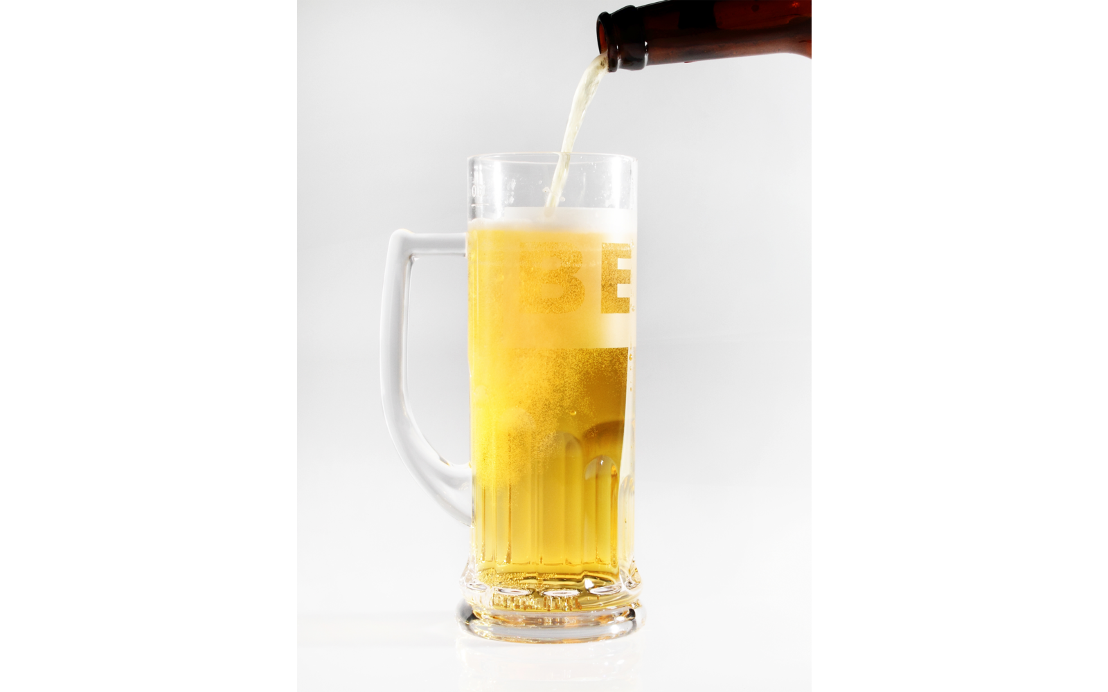 Bouteille brune verse une bière dorée dans une chope en verre transparente avec anse, mousse dessus, fond blanc