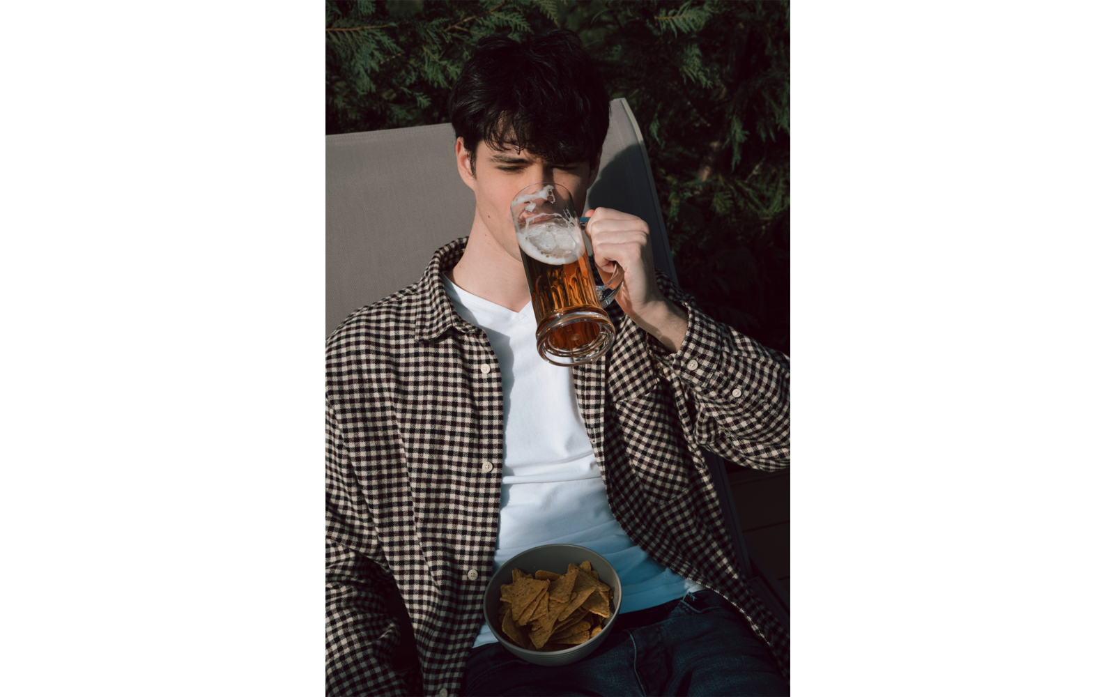 Jeune homme boit une bière dans une chope en verre, assis sur une chaise longue, porte une chemise à carreaux sur un T-shirt blanc, bol de chips tortilla sur les genoux, végétation verte en arrière-plan