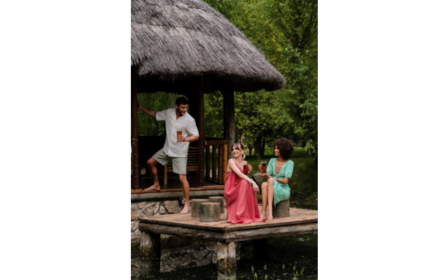 Trois personnes sur une jetée en bois et près d'une cabane au toit de chaume au bord d'un étang ; deux femmes sont assises sur la jetée en robes rose et verte tenant des boissons, l'une porte des lunettes de soleil ; un homme pieds nus se tient à l'entrée de la cabane avec une boisson