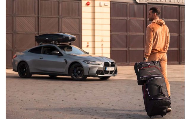 Mann in orangefarbener Freizeitkleidung mit Reisetasche und Rollkoffer steht neben einem silbergrauen Auto mit Dachbox vor geschlossenen Garagentoren