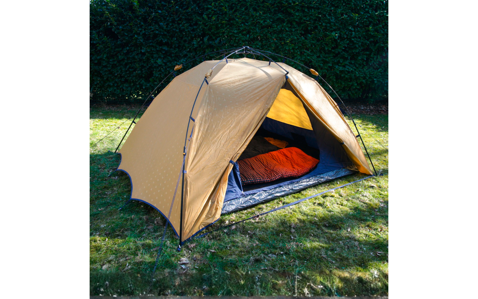 Beiges Zelt auf Rasen, geöffneter Eingang mit sichtbarem roten Schlafsack auf blauer Isomatte; Hecke im Hintergrund