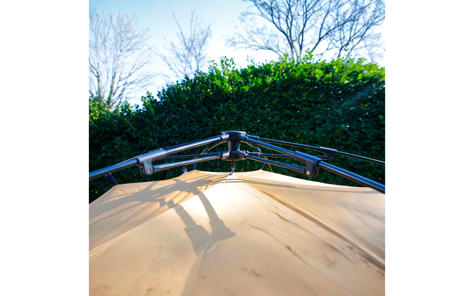 Blick nach oben auf eine hellbeige Stofffläche mit schwarzem Metallgestänge und Schatten darauf; im Hintergrund eine grüne Hecke, kahle Bäume und blauer Himmel