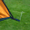Ecke eines orangefarbenen Zeltes mit schwarzem Gestänge, blauem Spannriemen und einem in grünen Rasen eingeschlagenen Metallhering