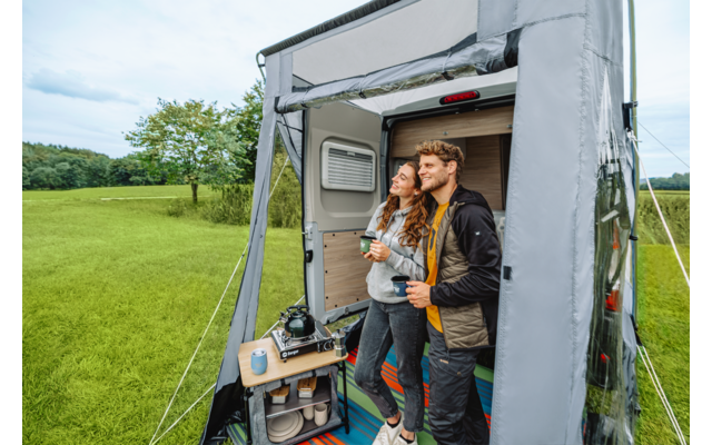 Zwei Personen stehen im Vorzelt eines Wohnmobils, halten jeweils eine Tasse und blicken nach rechts. Im Vorzelt ein Teppich und ein kleiner Tisch mit Kamera und weiterer Tasse; im Hintergrund Wiese und Bäume