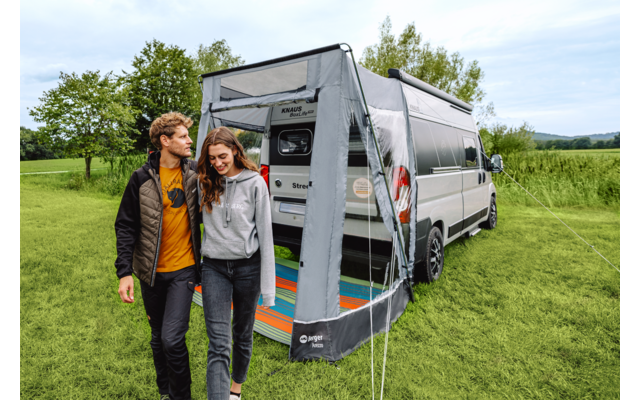Zwei Personen gehen nebeneinander vor einem Wohnmobil mit angebautem Vorzelt auf einer Wiese; im geöffneten Vorzelt liegt eine bunte Matte; Bäume und Himmel im Hintergrund