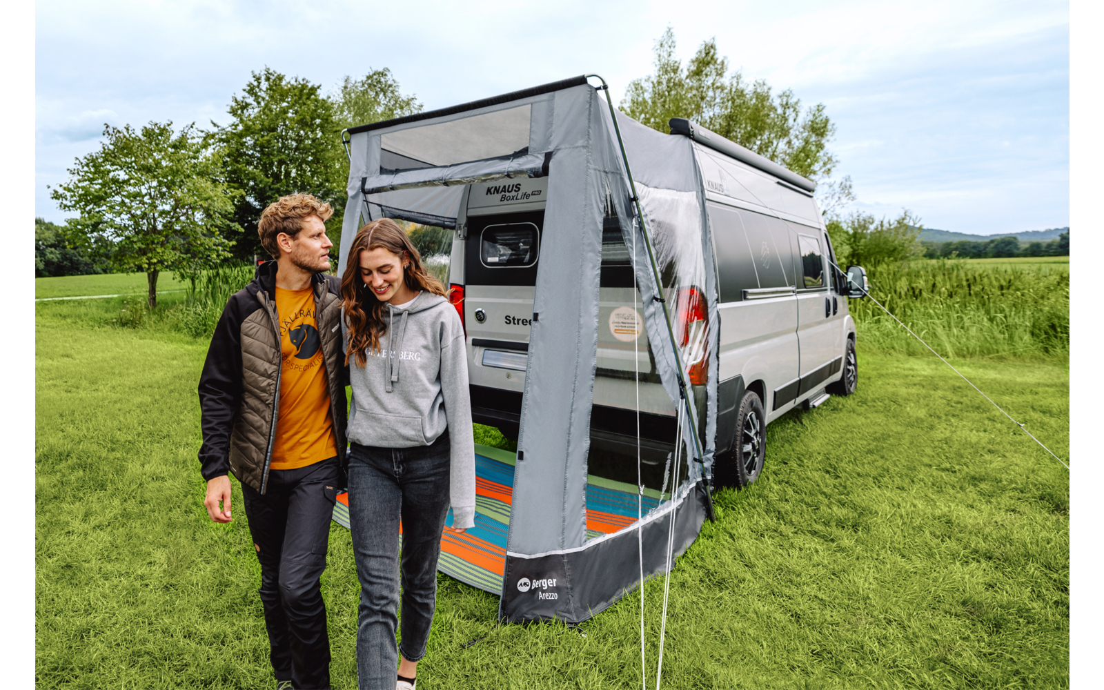 Zwei Personen gehen nebeneinander vor einem Wohnmobil mit angebautem Vorzelt auf einer Wiese; im geöffneten Vorzelt liegt eine bunte Matte; Bäume und Himmel im Hintergrund
