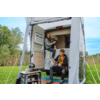 Open camper met voortent; een vrouw zit in de deuropening en houdt een thermosfles vast; een man zit op de treeplank en houdt een mok vast; campingtafel met gasstel en keukenspullen ervoor; grasveld en bomen op de achtergrond