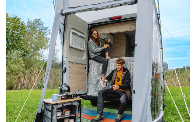 Open camper met voortent; een vrouw zit in de deuropening en houdt een thermosfles vast; een man zit op de treeplank en houdt een mok vast; campingtafel met gasstel en keukenspullen ervoor; grasveld en bomen op de achtergrond