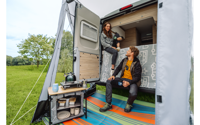 Twee personen in een open camper: één zit op het achterbed met een mok, de ander zit bij de instap met een mok; klein kampeertafeltje met kooktoestel en servies; kleurrijke vloermat; grasveld op de achtergrond