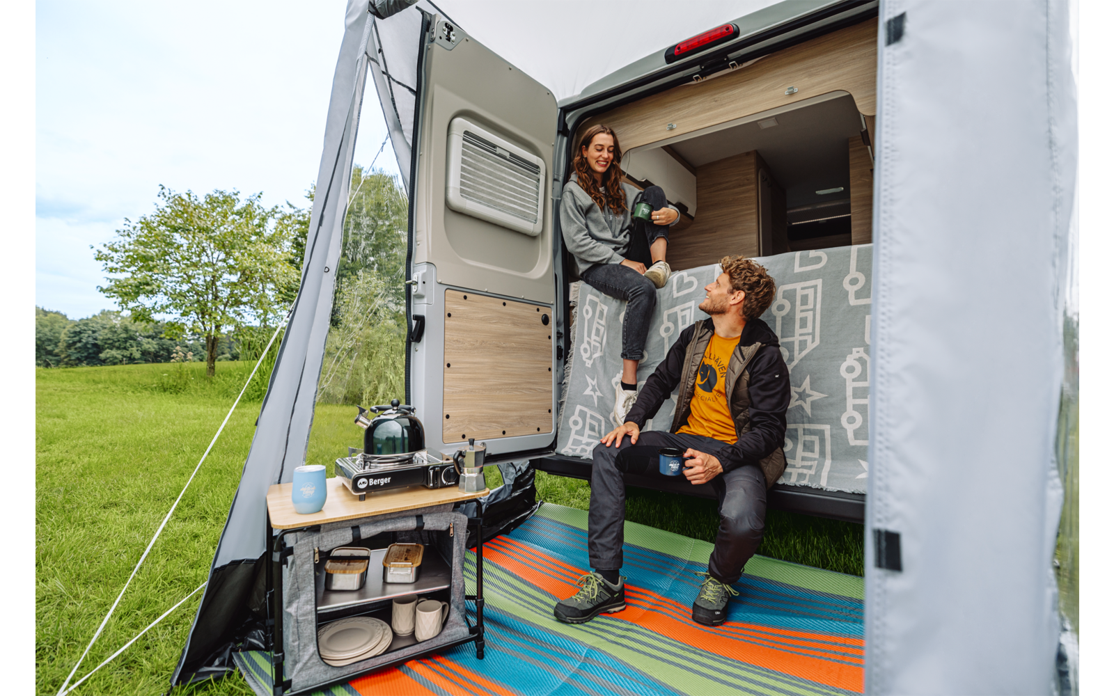 Twee personen in een open camper: één zit op het achterbed met een mok, de ander zit bij de instap met een mok; klein kampeertafeltje met kooktoestel en servies; kleurrijke vloermat; grasveld op de achtergrond