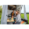 Twee volwassenen in het open voorbouw van een camper: een vrouw zit aan een opvouwbare tafel met een mok, een man zit achter haar; een campingkooktoestel en een groene thermocup op de tafel; grasveld en bomen op de achtergrond