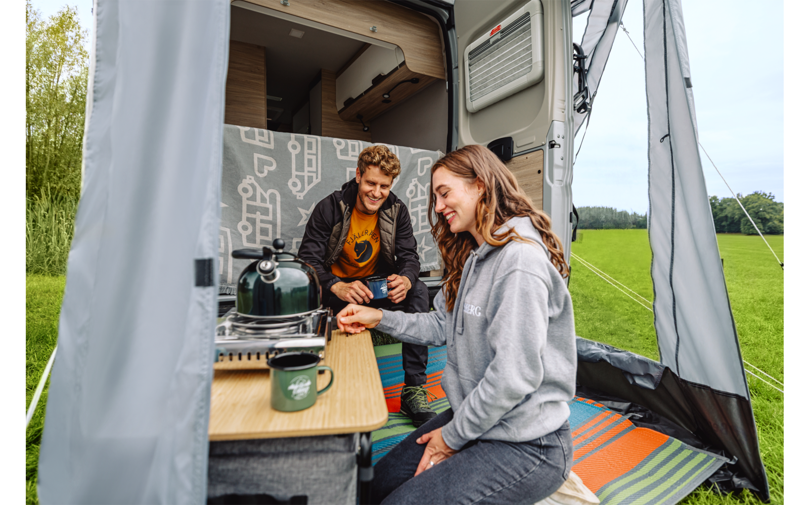 Twee volwassenen in het open voorbouw van een camper: een vrouw zit aan een opvouwbare tafel met een mok, een man zit achter haar; een campingkooktoestel en een groene thermocup op de tafel; grasveld en bomen op de achtergrond