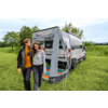 Twee personen lopen voor een geparkeerde witte camper met een aangebouwde luifel op een grasveld; bomen en een bewolkte lucht op de achtergrond