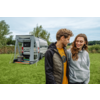 Twee glimlachende personen staan op gras voor een grijze camperbus met uitgeklapte luifel; naast de bus ligt een gestreepte mat en een draagbaar apparaat, op de achtergrond bomen en een bewolkte lucht