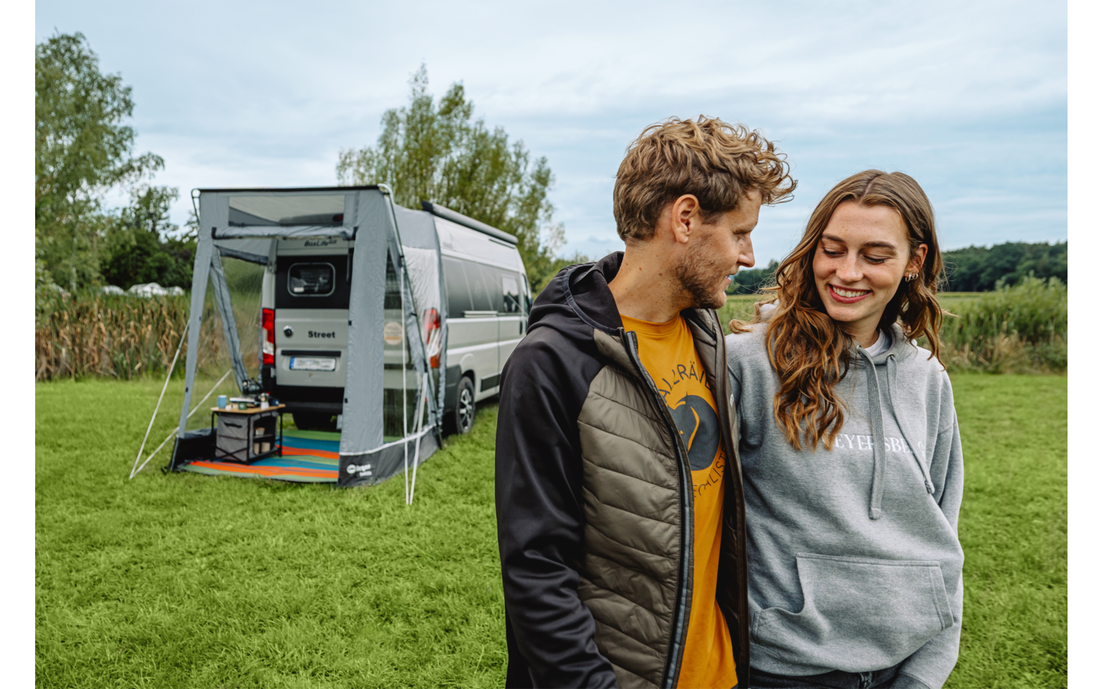 Twee glimlachende personen staan op gras voor een grijze camperbus met uitgeklapte luifel; naast de bus ligt een gestreepte mat en een draagbaar apparaat, op de achtergrond bomen en een bewolkte lucht